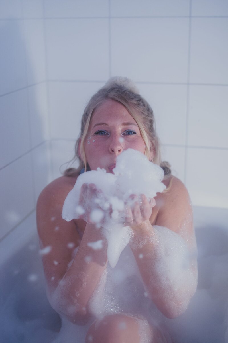 Blonde woman sits in bathtub and playfully flows suds at the camera.