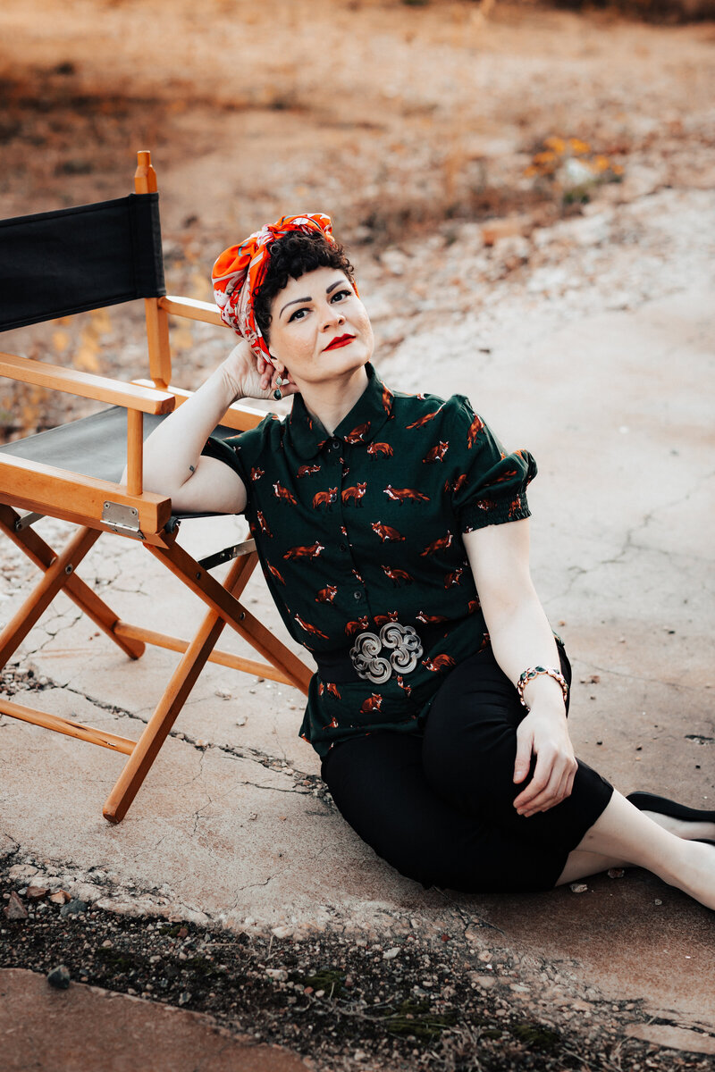 Laura is seated on a cement path outside, leaning against a director-style chair.  She is wearing a dark green, button-down shirt with orange foxes, as well as a red-patterned hair wrap and black cropped pants. Laura props her head up with her right hand, and looks calmly toward the viewer.