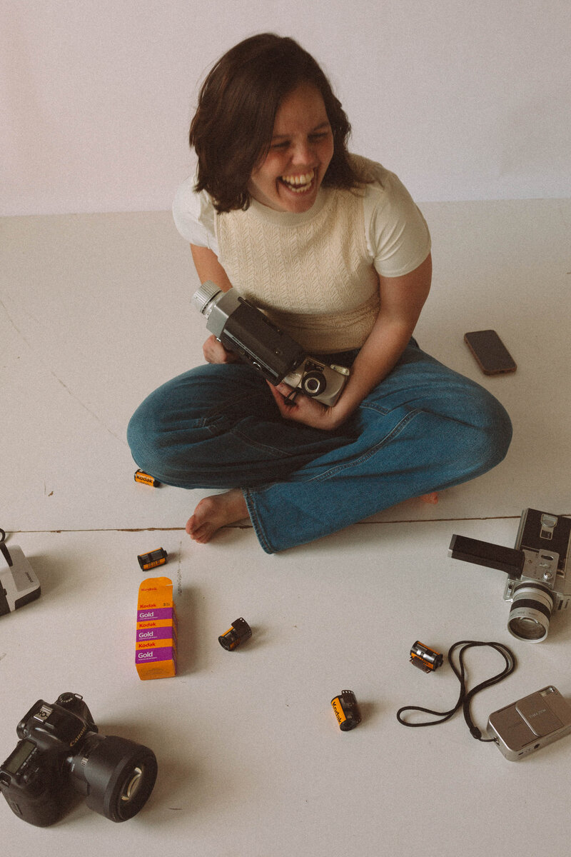 Photographer crouched down with elbows on knees, looking off thoughtfully, surrounded by camera gear and film.