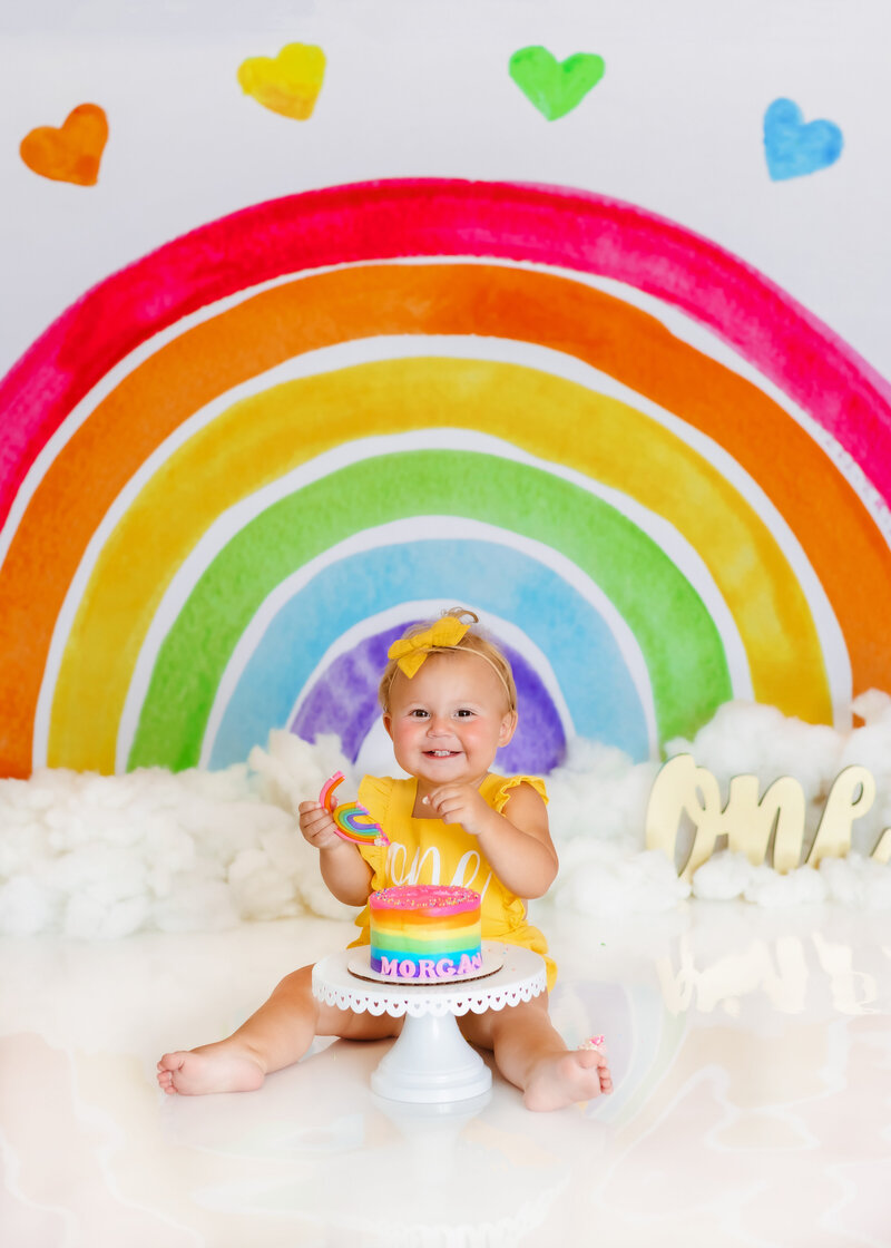 Family Photographer, a baby smiles with rainbow cake before her and a rainbow backdrop