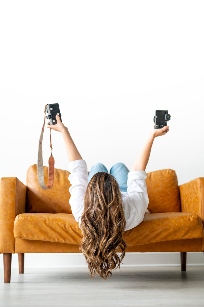 A fun shot of Kala laying on a large caramel colored chair with her back to the camera so her hair is dangling off the chair. In each hand she is holding a camera and raising them above her head.
