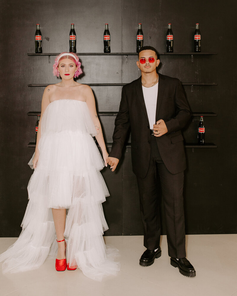 Trendy bride and groom stand in front of a display of coke bottles