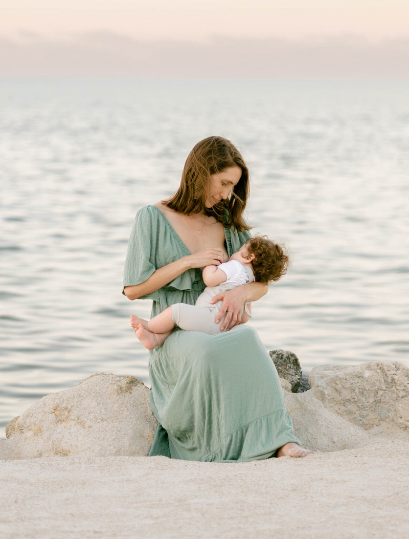 Mother lovingly holding and nursing her baby at sunset by the beach in Miami, Florida, captured by Claudia Amalia. Claudia is a wedding and lifestyle photographer based in Miami and Florida Keys, South Florida. She offers beautiful lifestyle photography packages and specializes in destination weddings, providing memorable and heartfelt photography services.