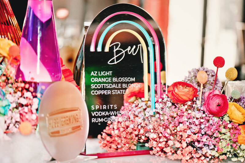 Neon-accented drink menu surrounded by colorful florals—photographed by MISGIF at a Phoenix corporate event to showcase curated bar details and sponsor branding.