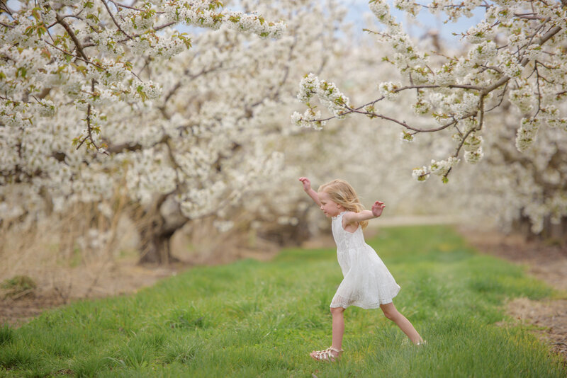 little girl in a white dress stomping through a flowering orchard with her arms swinging during photography with boise photographer Tiffany Hix in Boise Idaho