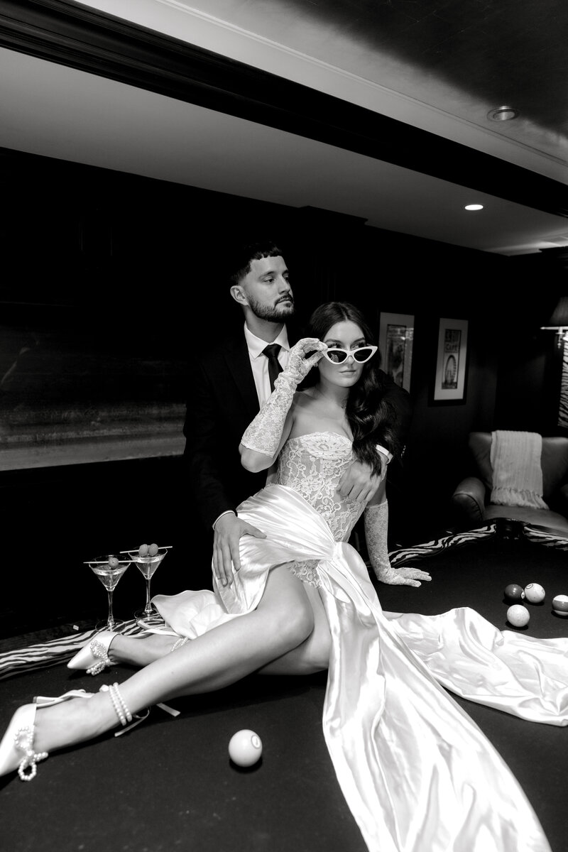 Newport Rhode Island Wedding Photographers | Rhode Island Caprio Captured Photography | Bride Wearing A White Silk Dress With Sunglasses Sitting On Pool Table While While Groom Is Holding Her