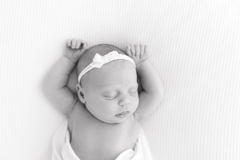 A black and white image of a baby girl laying on a white blanket with her arms above her head sleeping.