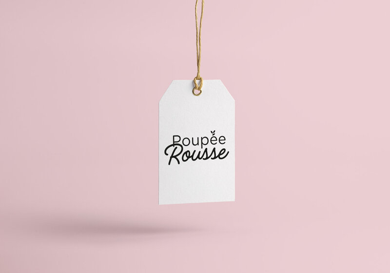 Étiquette blanche suspendue avec le logo “Poupée Rousse” sur fond rose poudré, réalisée par Studio Helloday. Image douce et élégante illustrant une création d’identité visuelle.