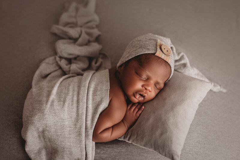 Newborn baby boy asleep, laying on mini gray pillow, gray fabric backdrop, laying on tummy with gray fabric draped over his diaper and wearing a gray sleepy cap