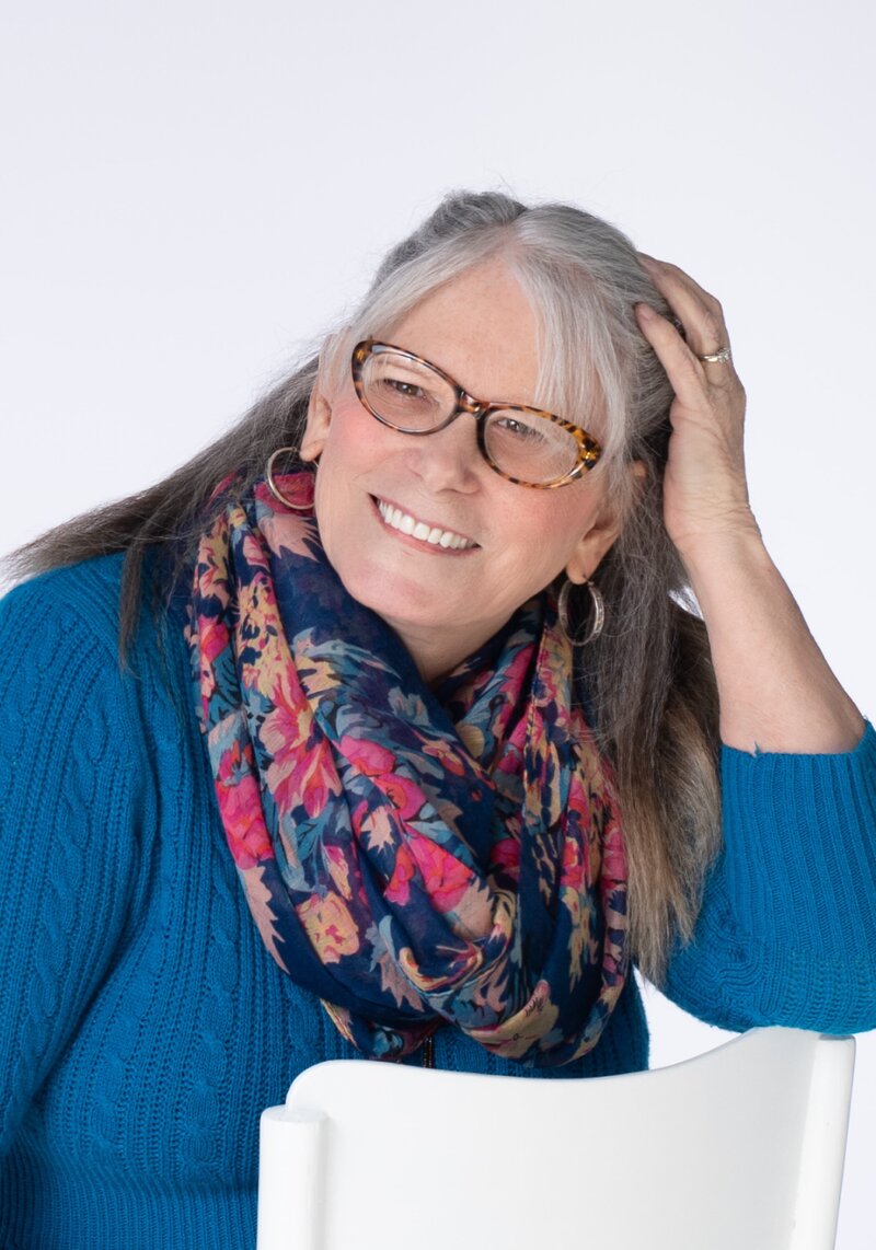 Image of a woman with grey hair wearing a blue sweater and floral scarf with glasses and smiling