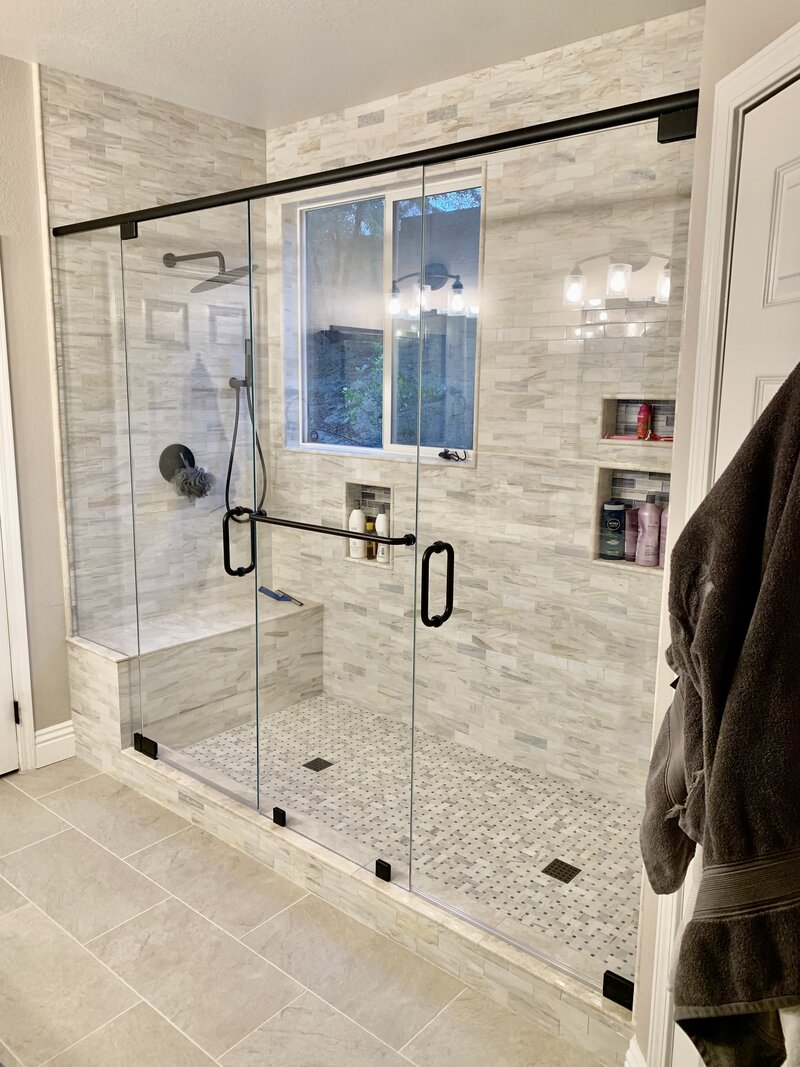 A newly remodeled shower with two glass doors and black hardware. The shower is tiled with black and grey horizontal tiles and there is a seat at the back.