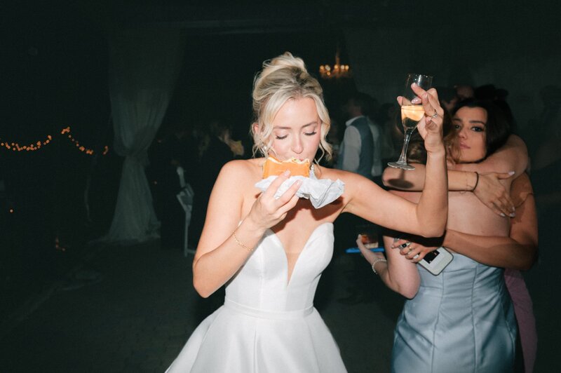 A bride in her wedding gown enjoys a snack while holding a glass of champagne during a fun and casual reception moment. 	9.	Ninth Image (Veil Portrait)