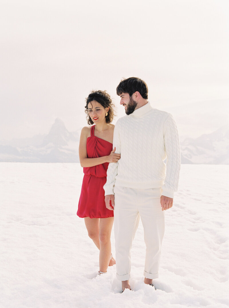 Woman in red dress holds husbands arm as they walk through the snow
