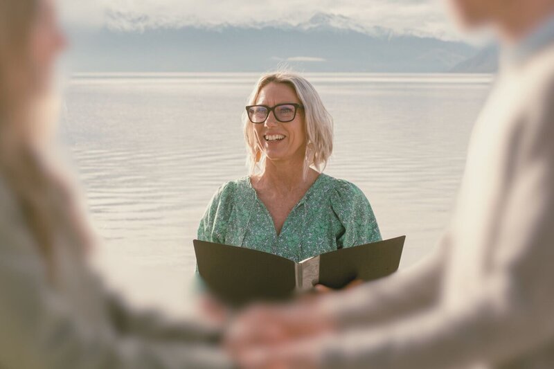 Close up of celebrant Kirsty performing a wedding ceremony beside a beautiful lake with the snow-capped mountains in the distance. Kirsty is wearing black glasses and a bright green dress. The bride and groom are blurred in the foreground.