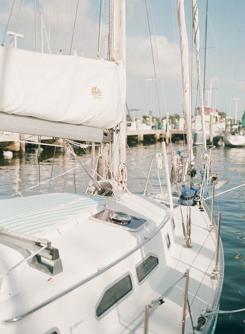 A sailboat is docked at a marina, with ropes and sails neatly arranged. The sunlight creates a soft reflection on the water, and other boats and a pier are visible in the background.