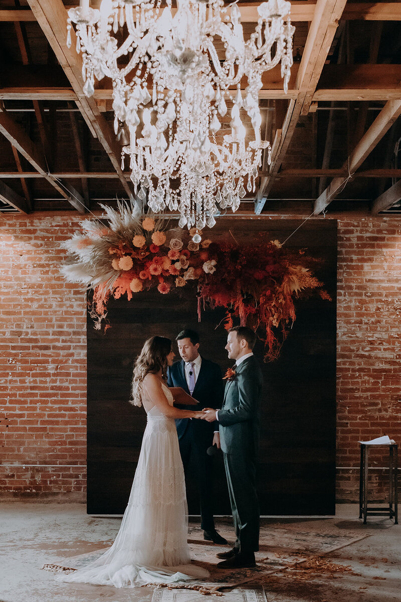 Wedding ceremony against walnut ceremony backdrop , chandeliers and  ruby colored florals