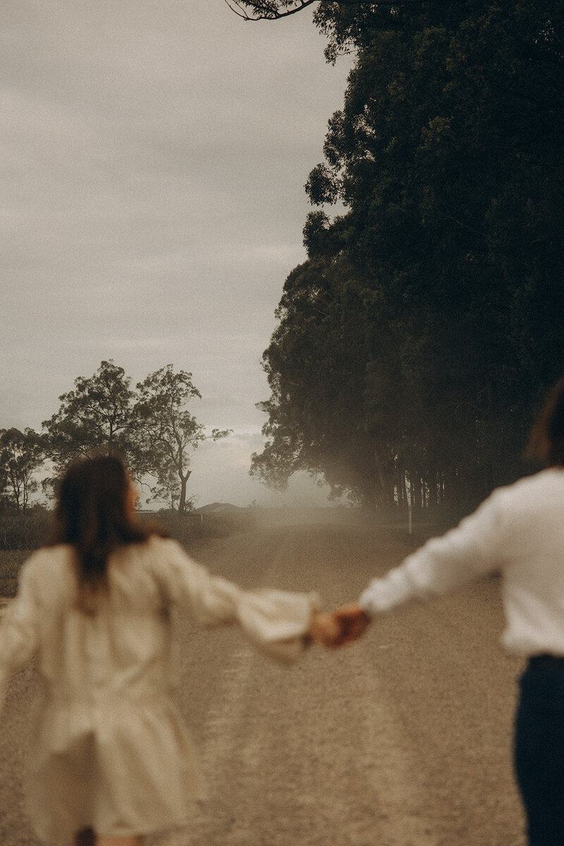 Adventurous couple holding hand in the foreground with a dirt road in the background taken on the Sunshine Coast by couples photographer