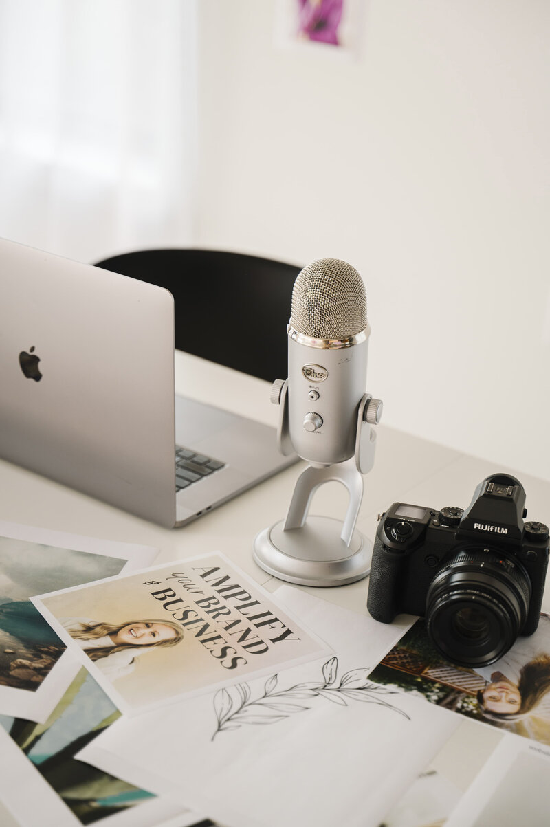 A white table with a laptop, podcast mic and a camera on top of printed brand photos taken by Krista Marie