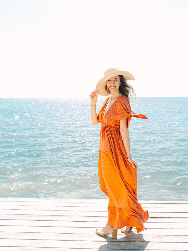 Kilee in an orange dress and straw hat sitting in chair on the beach with ocean behind her