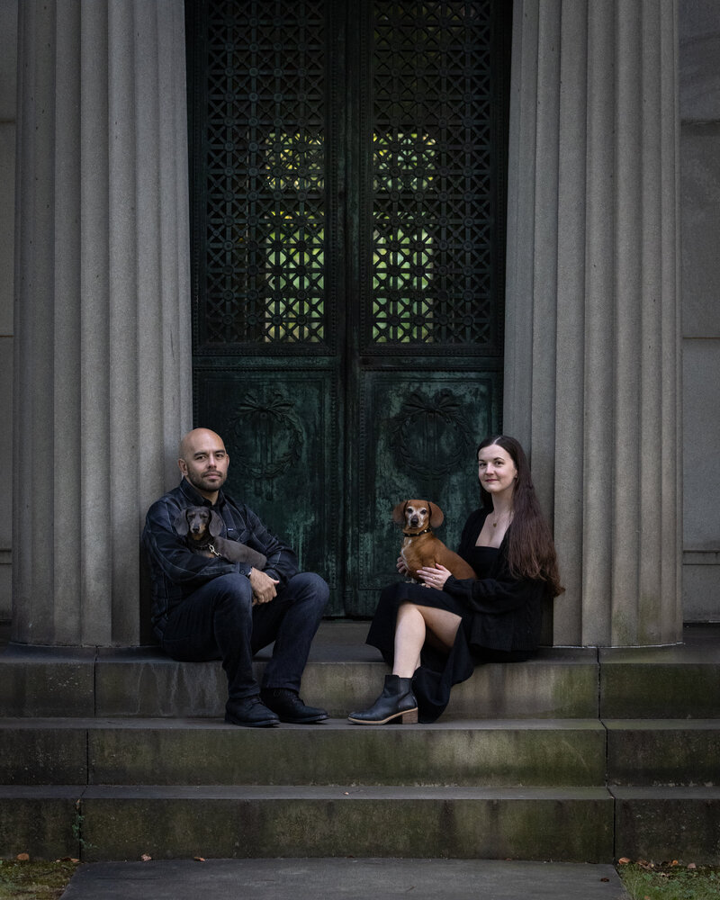 A man and a woman are sitting on steps of a mausoleum in Lakeview Cemetery in Cleveland Ohio. There are dachshunds on each of their laps.