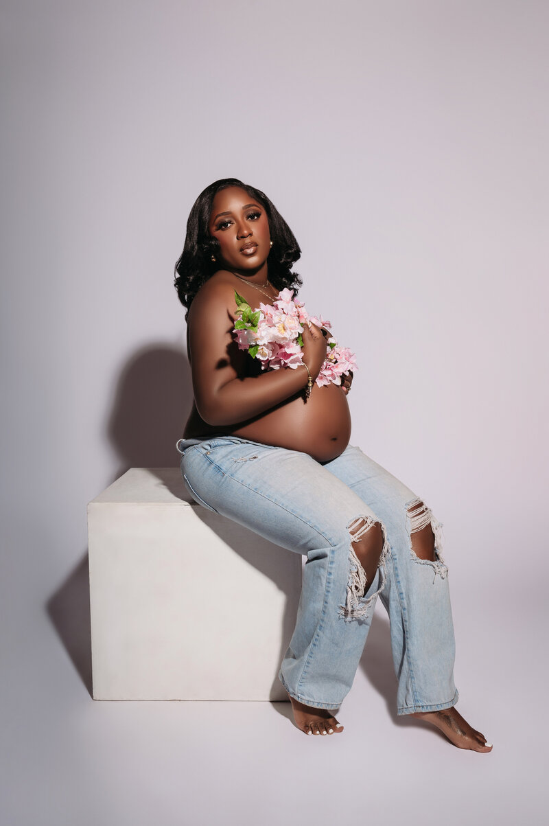 beautifl pregnant woman wearing jeans and holding flowers while sitting on white cube prop and posing into the camera