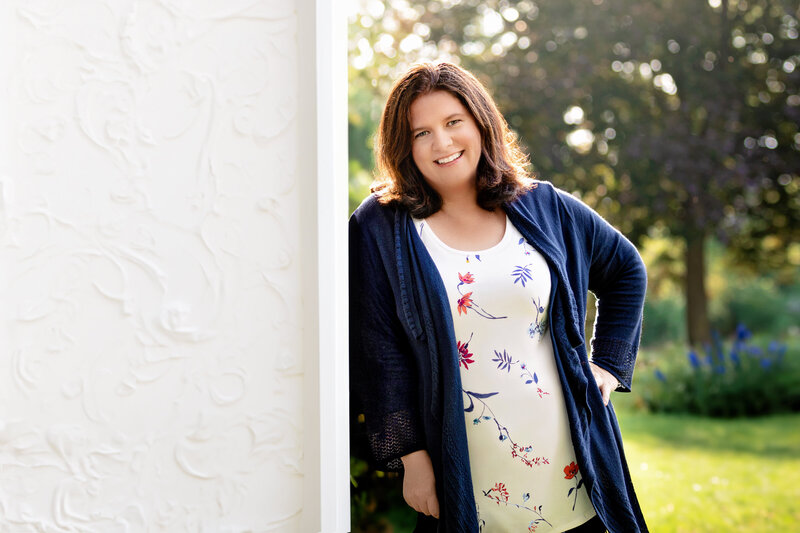 Smiling woman standing outdoors in soft sunlight, leaning against a white wall while wearing a navy cardigan and a floral top.