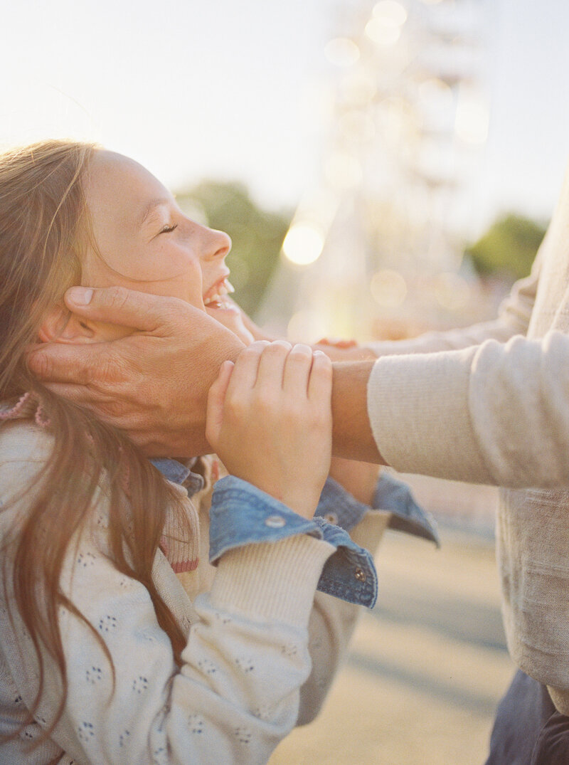 Tween daughter laughs joyously in a sun-soaked film photo as her father's hands embrace her face.