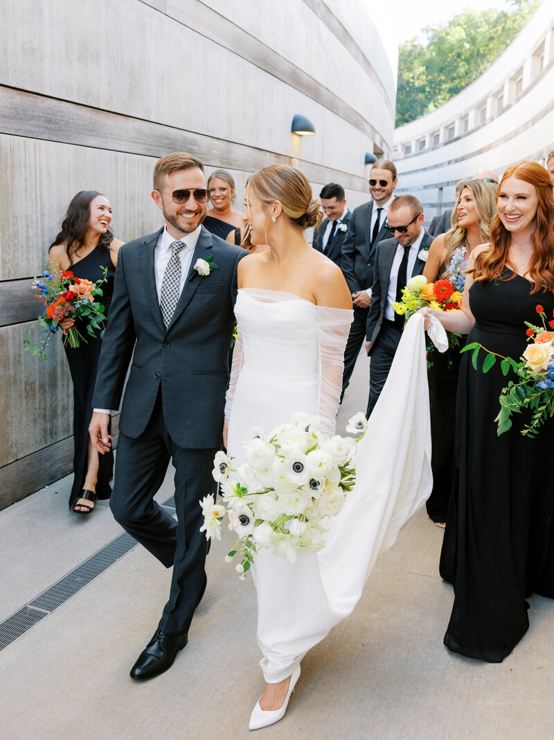 wedding party walks and laughs towards camera with black bridesmaids dresses, elegant attire, and colorful florals