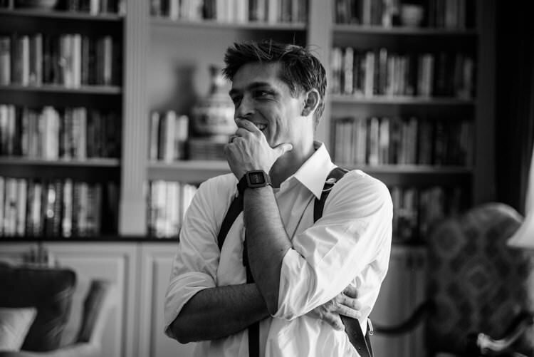 Black and white portrait of photographer Rob Futrell in white dress shirt and suspenders, hand to chin in thoughtful pose, standing in front of bookshelves during St. Augustine Courthouse wedding preparations