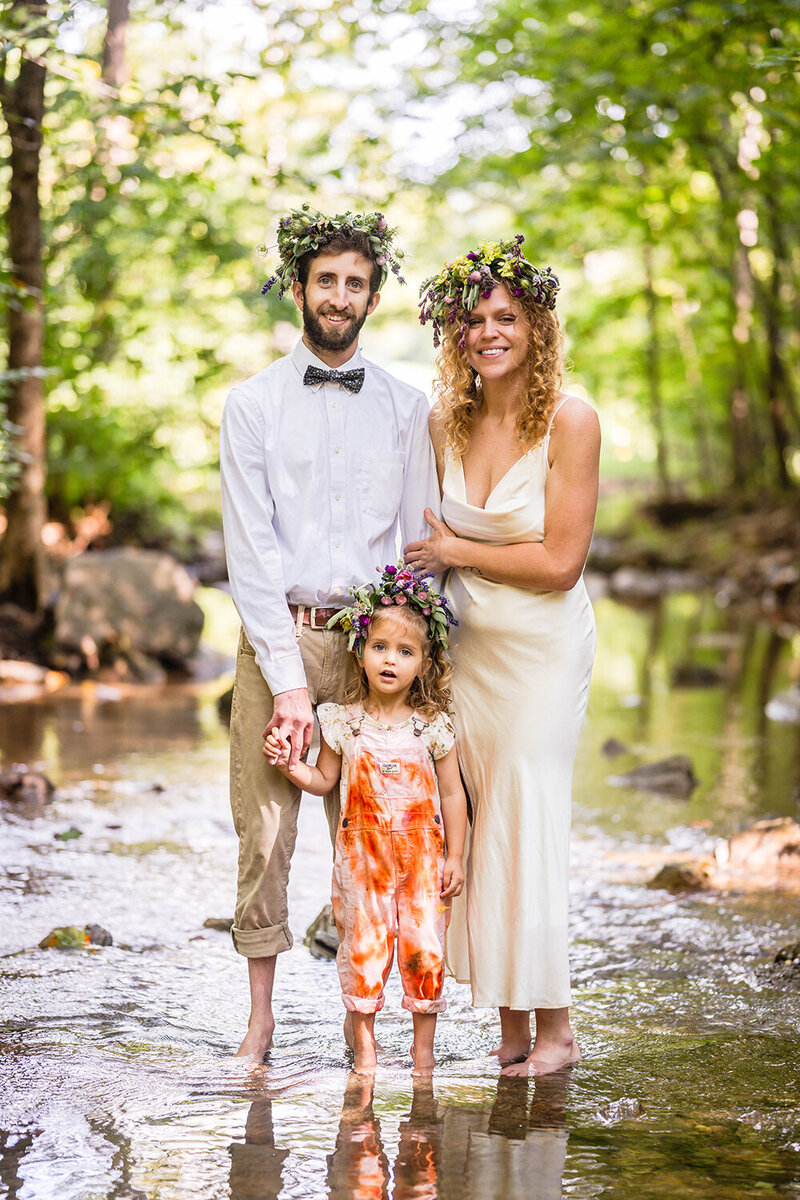 A man, a woman, and their child stand barefoot in a shallow creek and smile for a photo during their elopement day inRoanoke, Virginia at Fishburn Park.