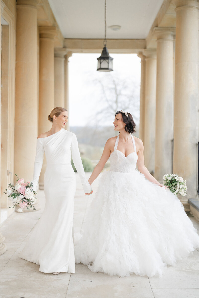 A stunning same sex wedding at Buxted Park, featuring two beautiful brides in elegant gowns. They walk hand in hand in the stately manor, beaming with love and joy. The scene is timeless, romantic, and full of pure happiness.