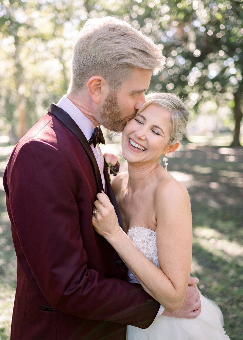 groom in burgandy tux kissing his bride on the cheek