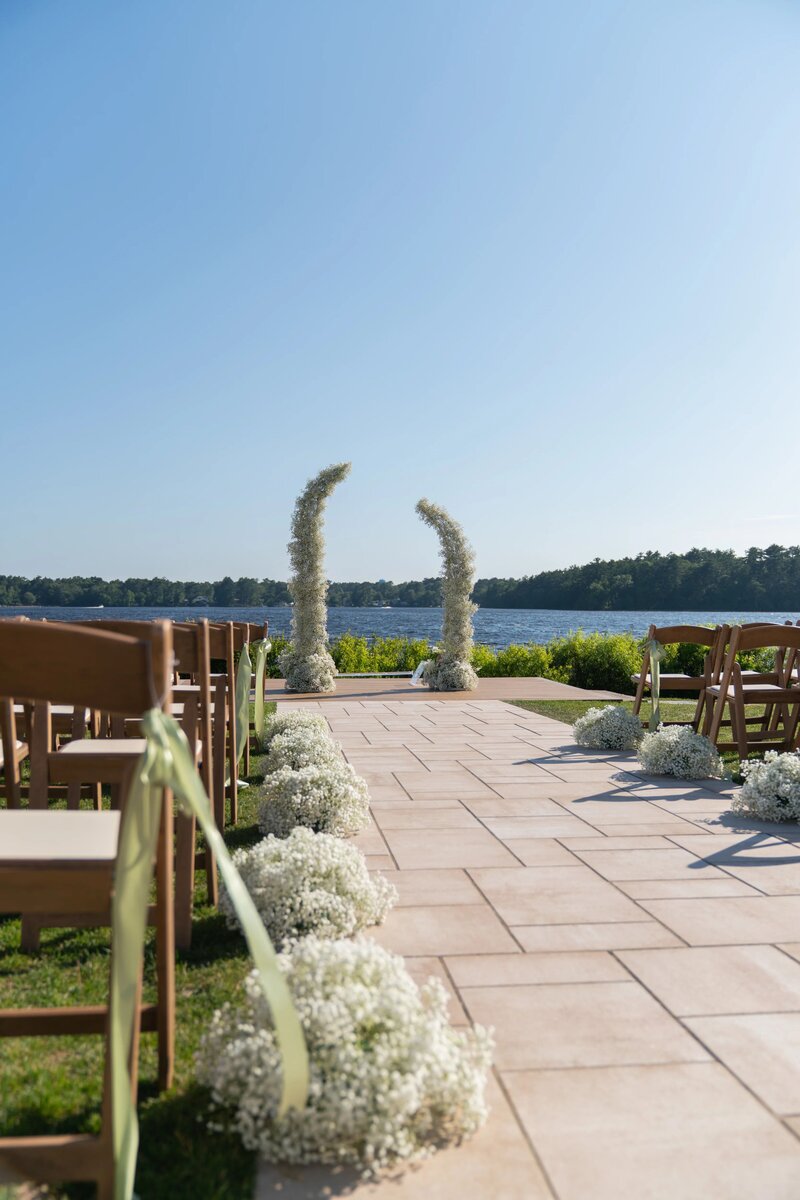 New England Wedding Photographer Pricing | Kelsey Sheehan Photography Timeless Rhode Island Weddings | Outdoor wedding aisle by a lake, lined with white flowers and wooden chairs. Floral arch at the end. Clear blue sky, peaceful and romantic setting.