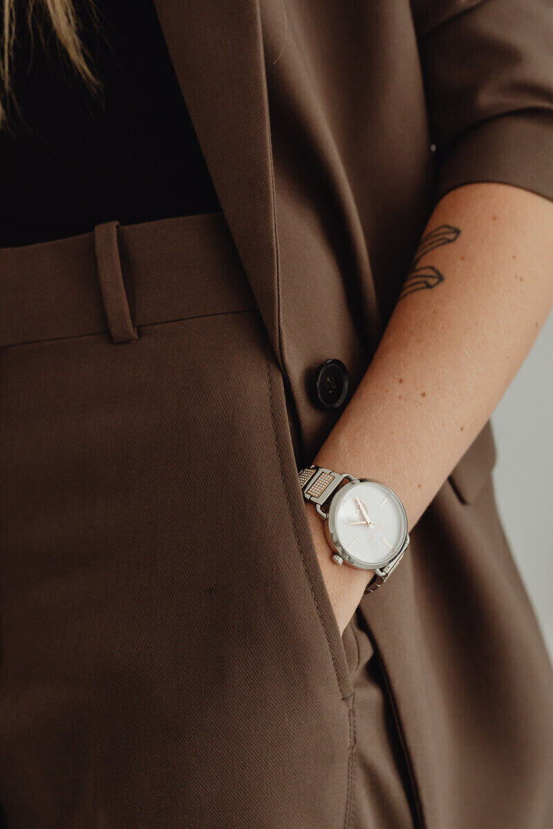 A woman in a brown suit, elegantly accessorized with a stylish watch, exuding professionalism and confidence.