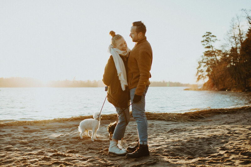 Engaged couple and their two dogs are on the beach in Espoo in Finland. The couple  is standing facing and holding each other and the woman is laughing.