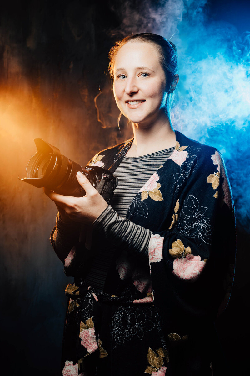 A photographer holding a camera and smiling with blue and orange smoke behind them 