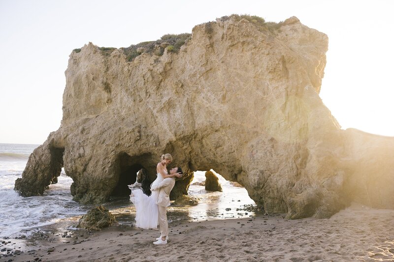 A groom picks up the bride in front of a big sea stack at El Matador State Beach.