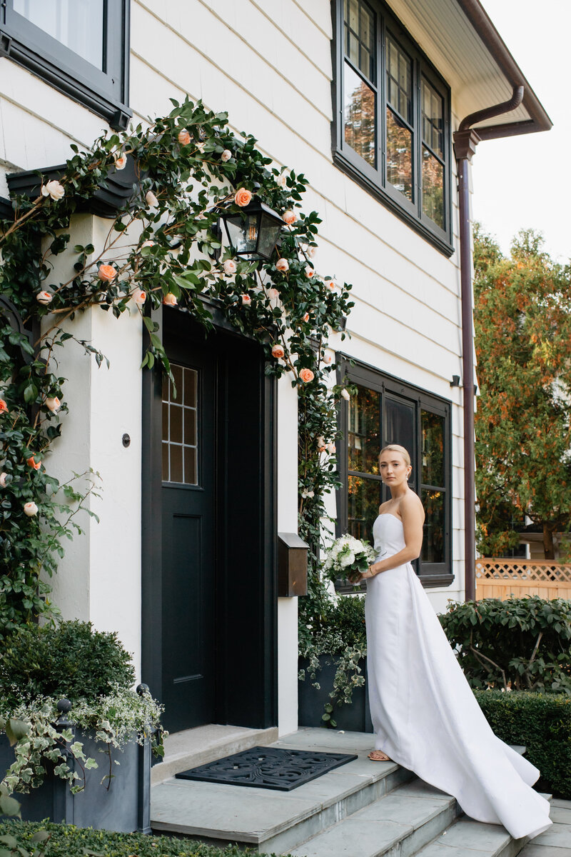 Bride in an elegant white gown standing by a floral-decorated doorway of a classic home, poised and graceful.