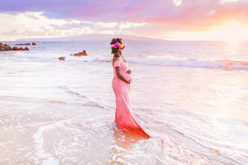 Pregnant woman in a long, pink, off-shoulder dress embraces her belly on the beach for her maternity photos