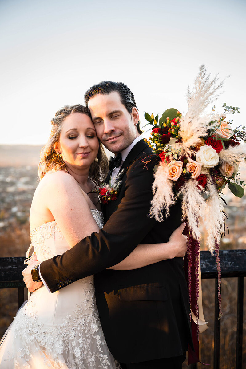 A couple embrace on their elopement day. They press their temples together and close their eyes as they take a breath during their elopement day at Mill Mountain Park at the Rockledge Overlook. A beautiful and large bouquet with long ribbons cascades behind the groom as the bride sets it on his back.