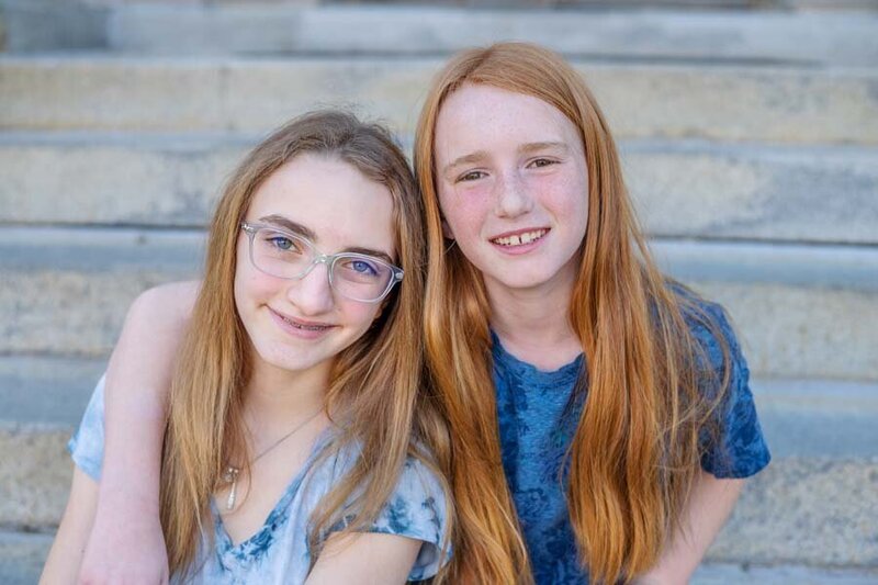 Two smiling young girls with glasses, one blonde and one redhead, sitting closely together on stone steps.