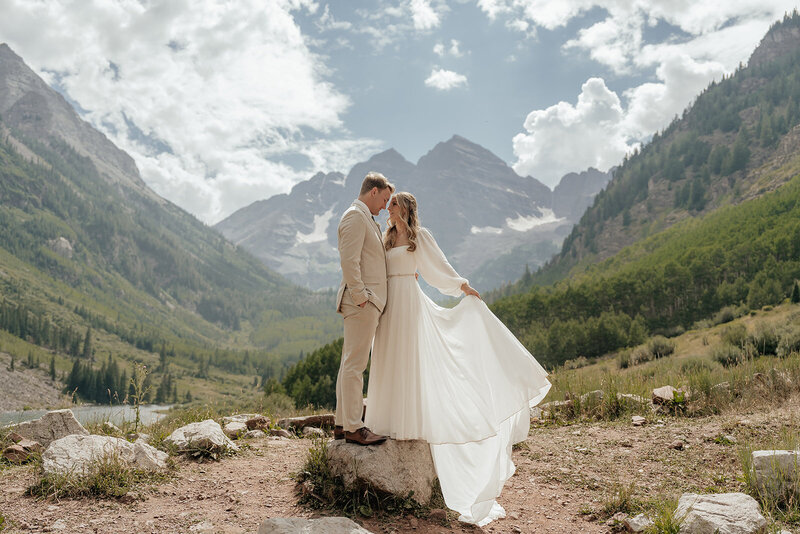 A couple shares an intimate moment surrounded by the towering peaks of Maroon Bells in Colorado, the bride’s elegant long-sleeved gown flowing in the breeze beneath a bright summer sky, captured by Sydney Breann Photography.