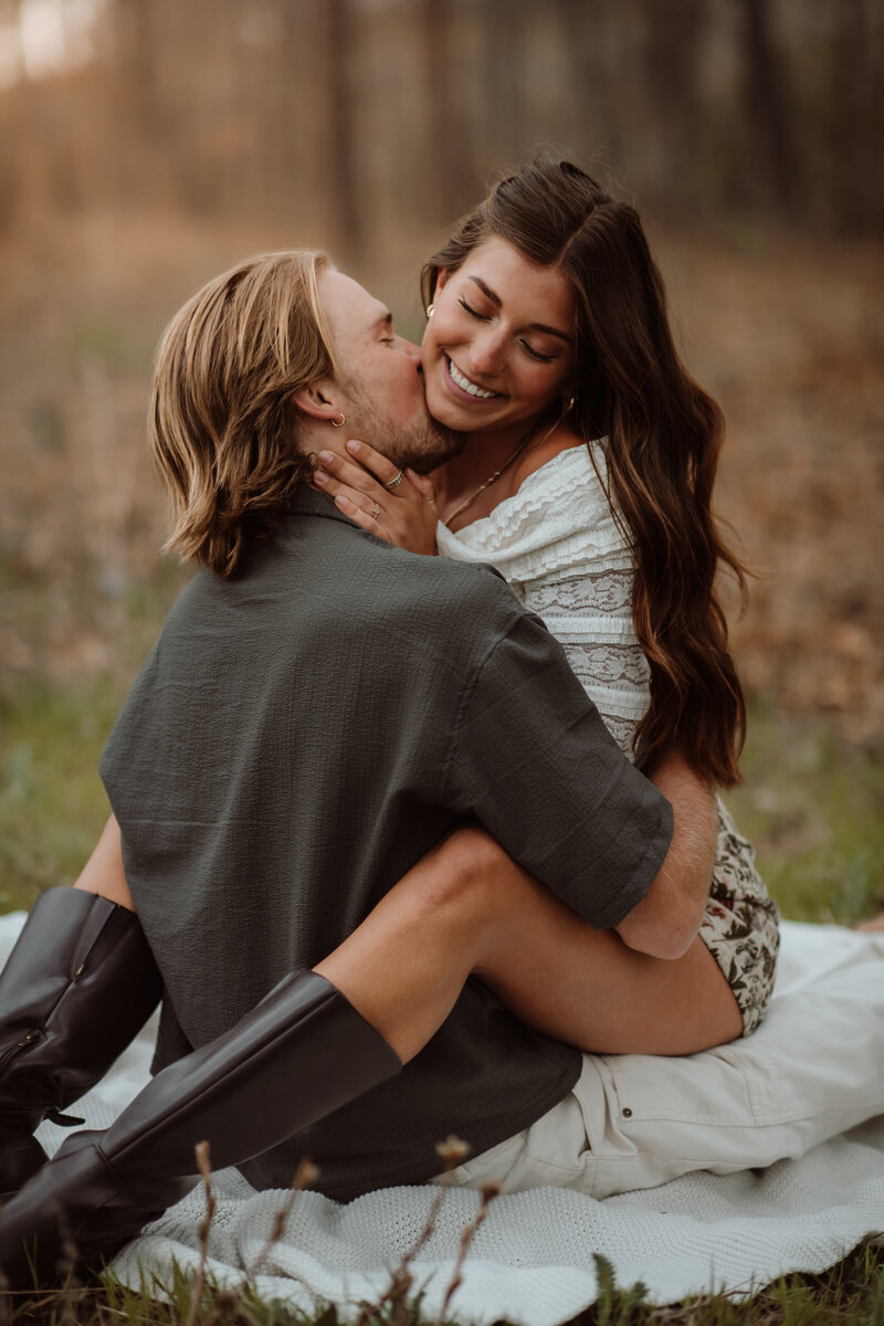 A woman sitting on her fiance's lap on a picnic blanket as he kisses her neck at Dawson Butte Open Space Colorado. 