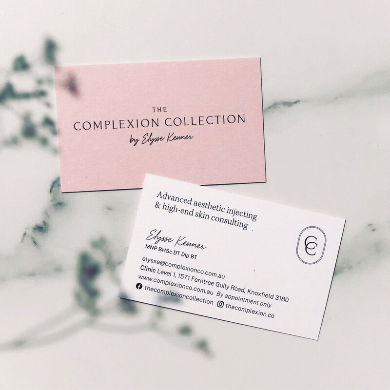 Flatlay of The Complexion Collection business cards showing front and back design, pastel pink and logo design on front and information on back sitting on a marble background with flower shadows overlayed