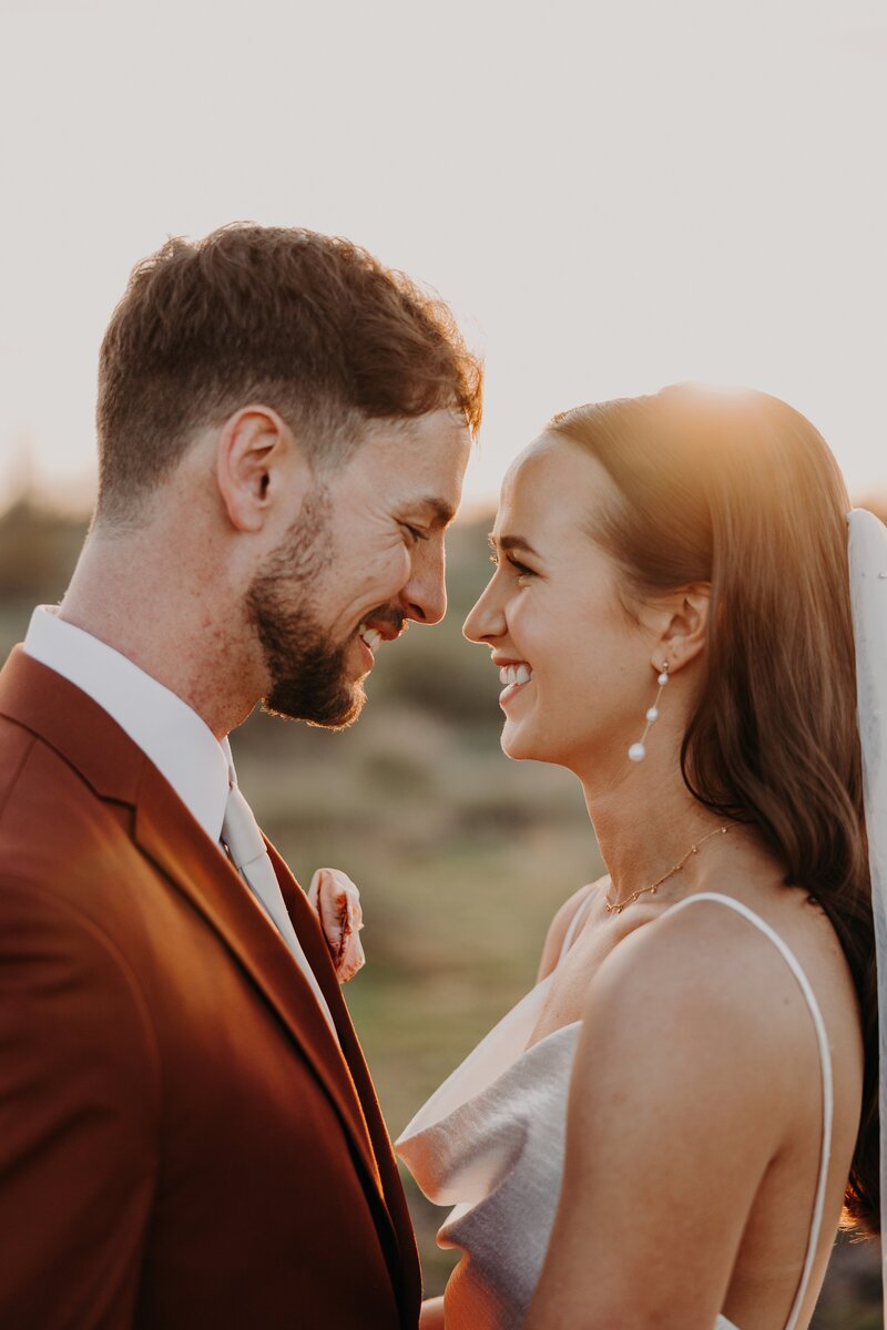 Bride and groom laugh together while almost kissing in the desert at sunset