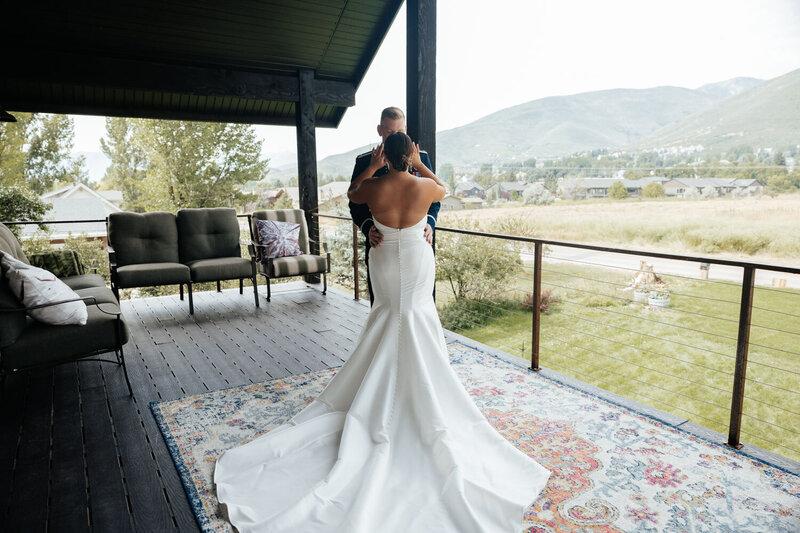 Bride holding the groom's face, standing on a deck with mountains in the background