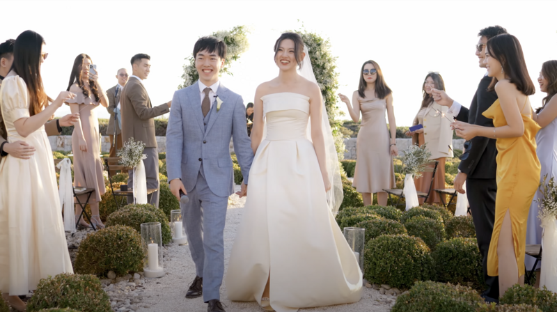 Newlyweds walking hand in hand as guests cheer during a romantic outdoor wedding in Greece, filmed in golden light with a soft, cinematic documentary wedding style