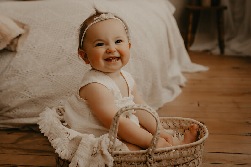 A one year old girl sitting in a moses basket while looking and smiling directly into the camera in a photography studio in Rochester, NY