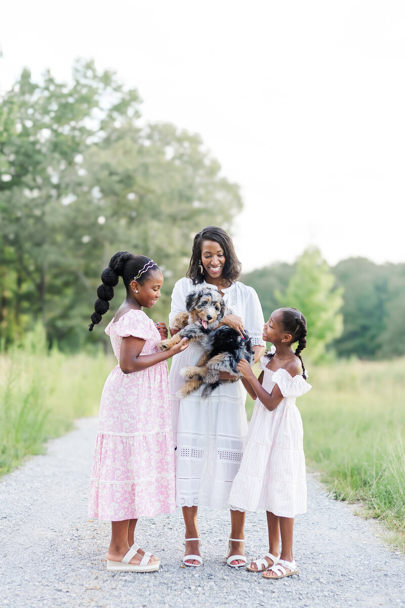 Mom holding puppy with her two daughters standing beside her all smiling down at puppy standing on gravel road at private property location in Wilsonville AL 