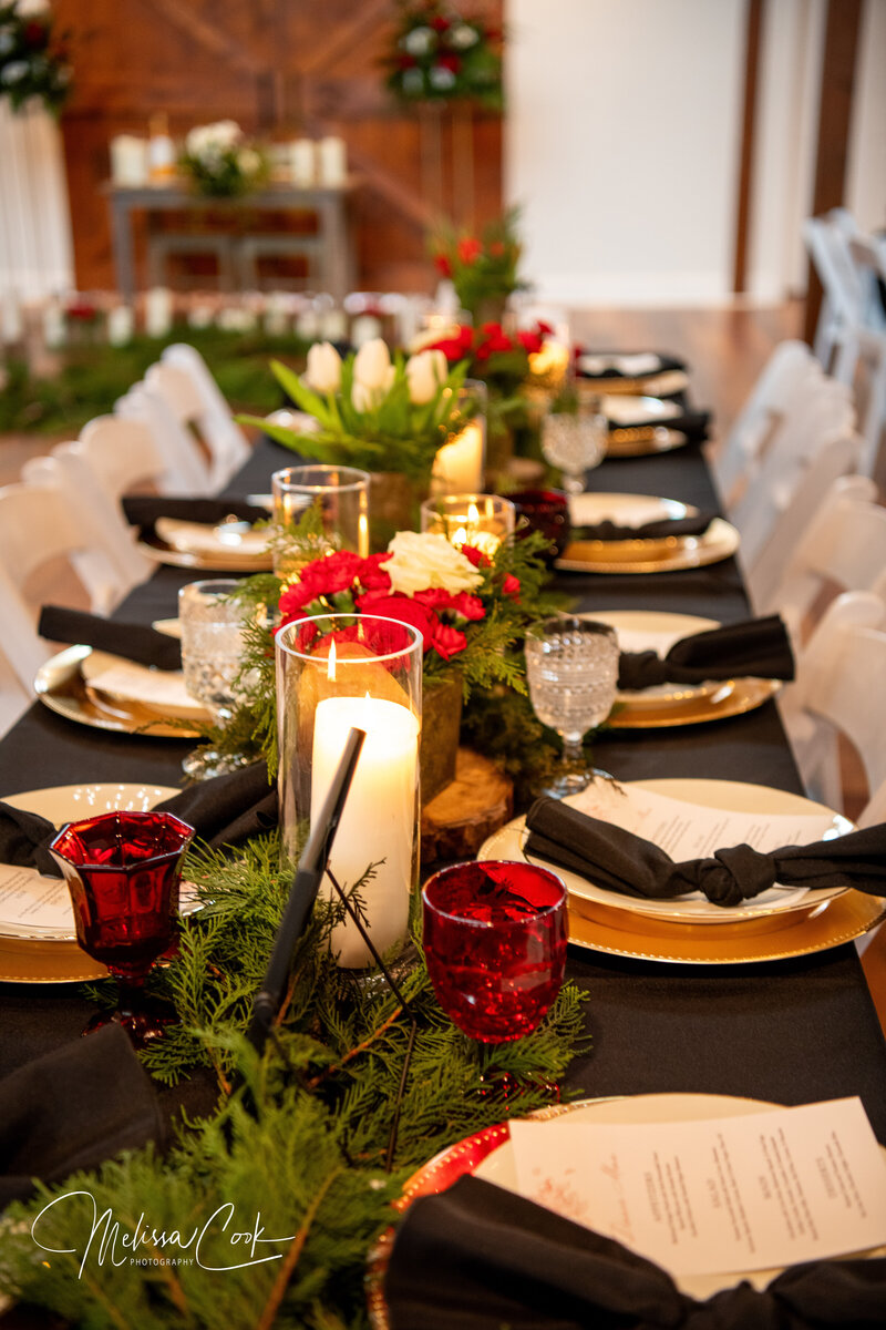 A festive dining table is set with black napkins, gold-rimmed plates, and red glassware. A centerpiece of greenery, candles, and red and white flowers runs along the table. White chairs surround the setup in a warmly lit room reminiscent of an elegant reception venue gallery.